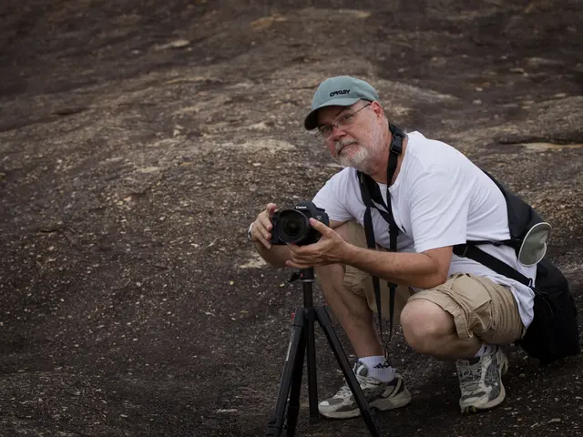 There is a old man in a squatting position on a rock. He is wearing a cap,spectacles,shoes,a white...